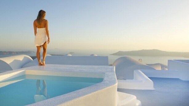 A woman standing by a pool with a view of the sea and islands. - Avista Suites (Santorini)