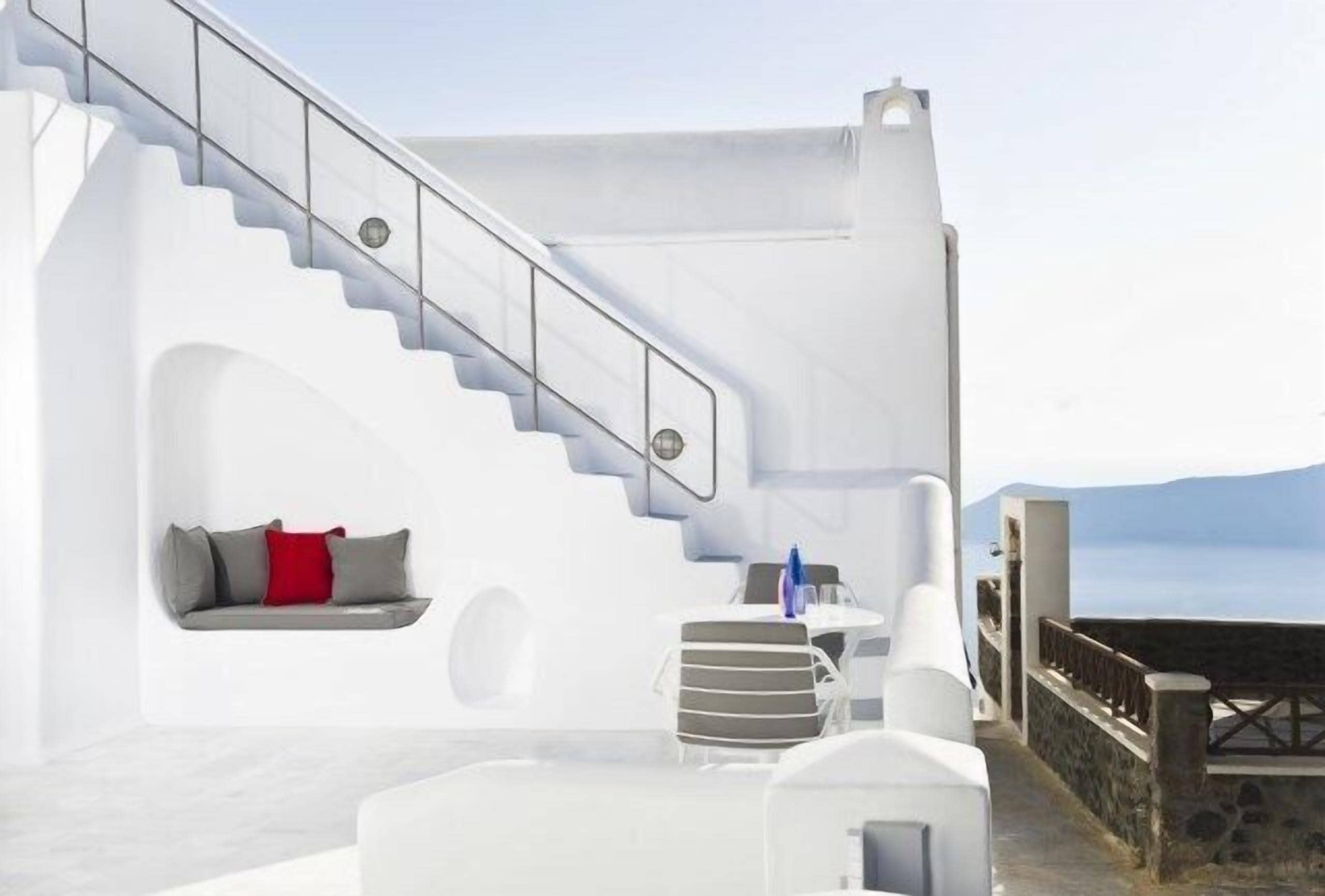 A modern white interior with a staircase, a seating area with cushions, and a view of the sea.