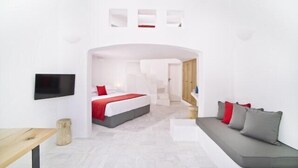 A modern hotel room with a bed, a sofa, a TV, and a small table. - Avista Suites (Santorini)