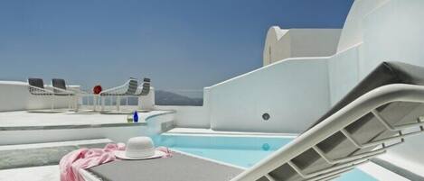 A rooftop pool area with lounge chairs and a table, clear skies, and a view of the sea.