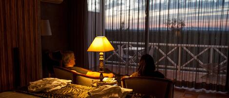 A hotel room with a bed, a lamp, a chair, and a view of the sunset through the window.