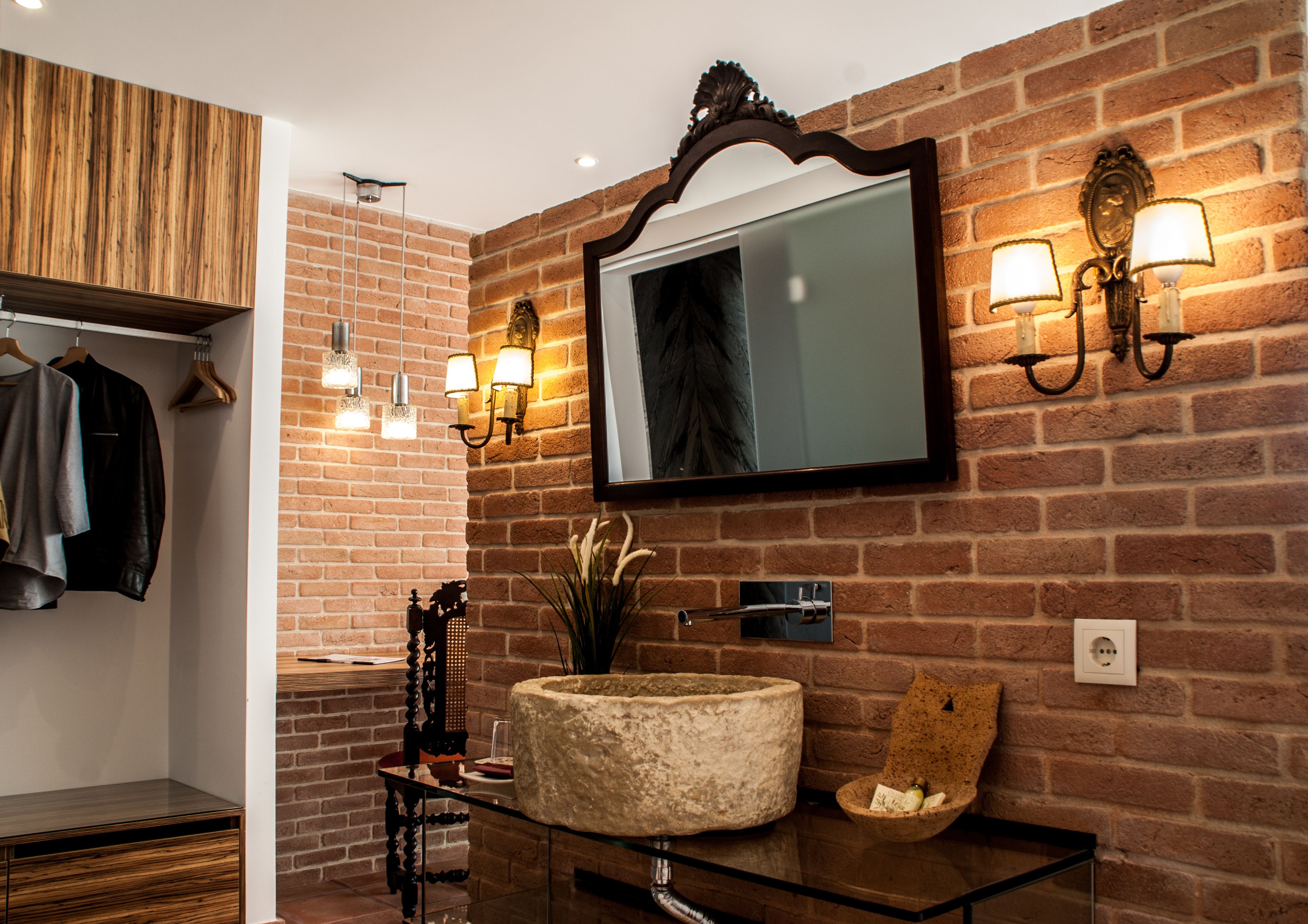 A bathroom with a brick wall, a wooden vanity, a mirror with wall sconces, and a glass sink.