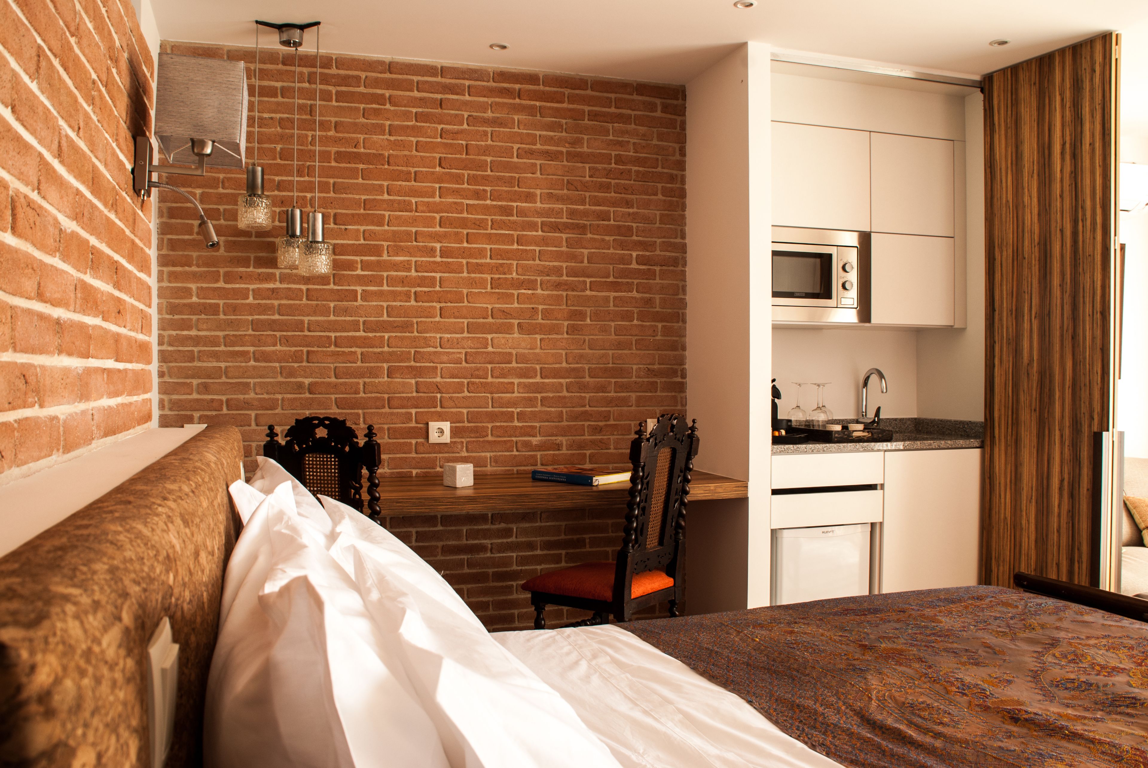 Una camera d'albergo con parete in mattoni, un letto con lenzuola bianche, un poggiatesta in legno, una scrivania con una sedia, un forno a microonde e un angolo cottura.
