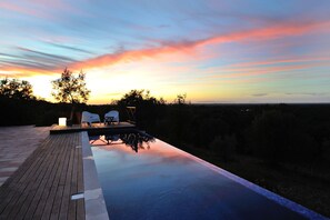 En infinitypool med ett trädäck och två vita stolar, med utsikt över ett landskap med träd och en solnedgång.