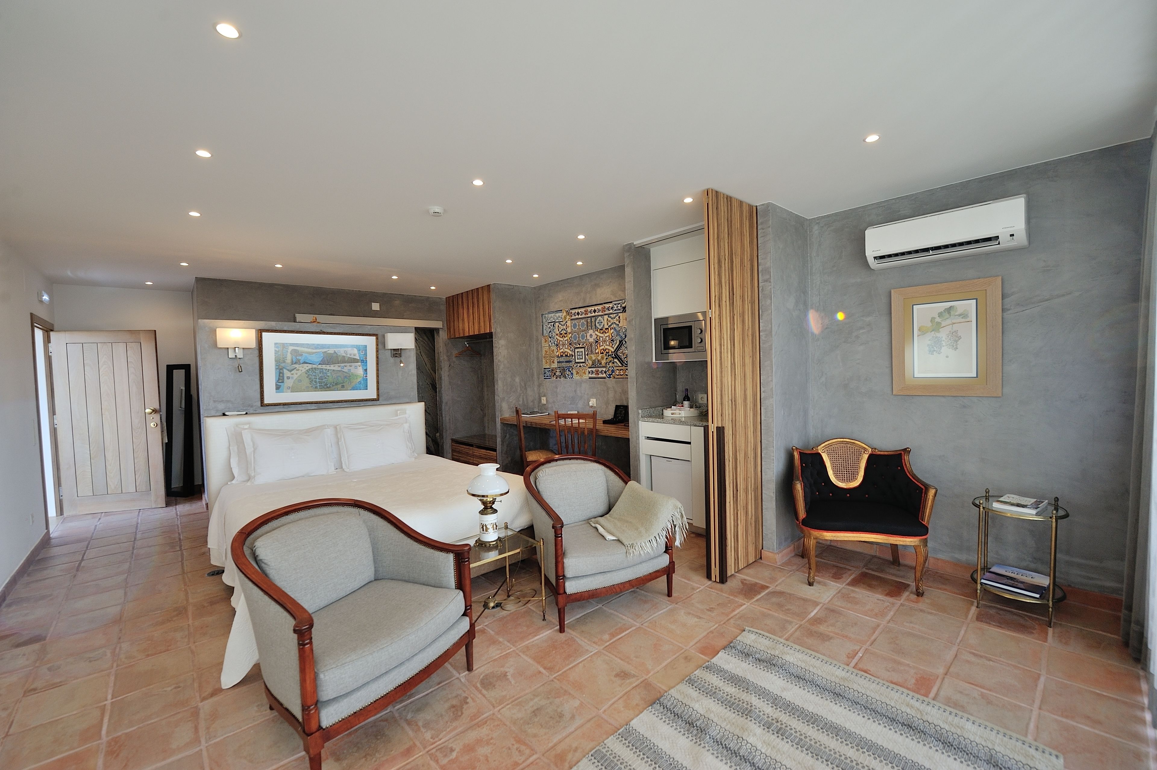 A hotel room with a bed, two armchairs, a desk, and an air conditioning unit.