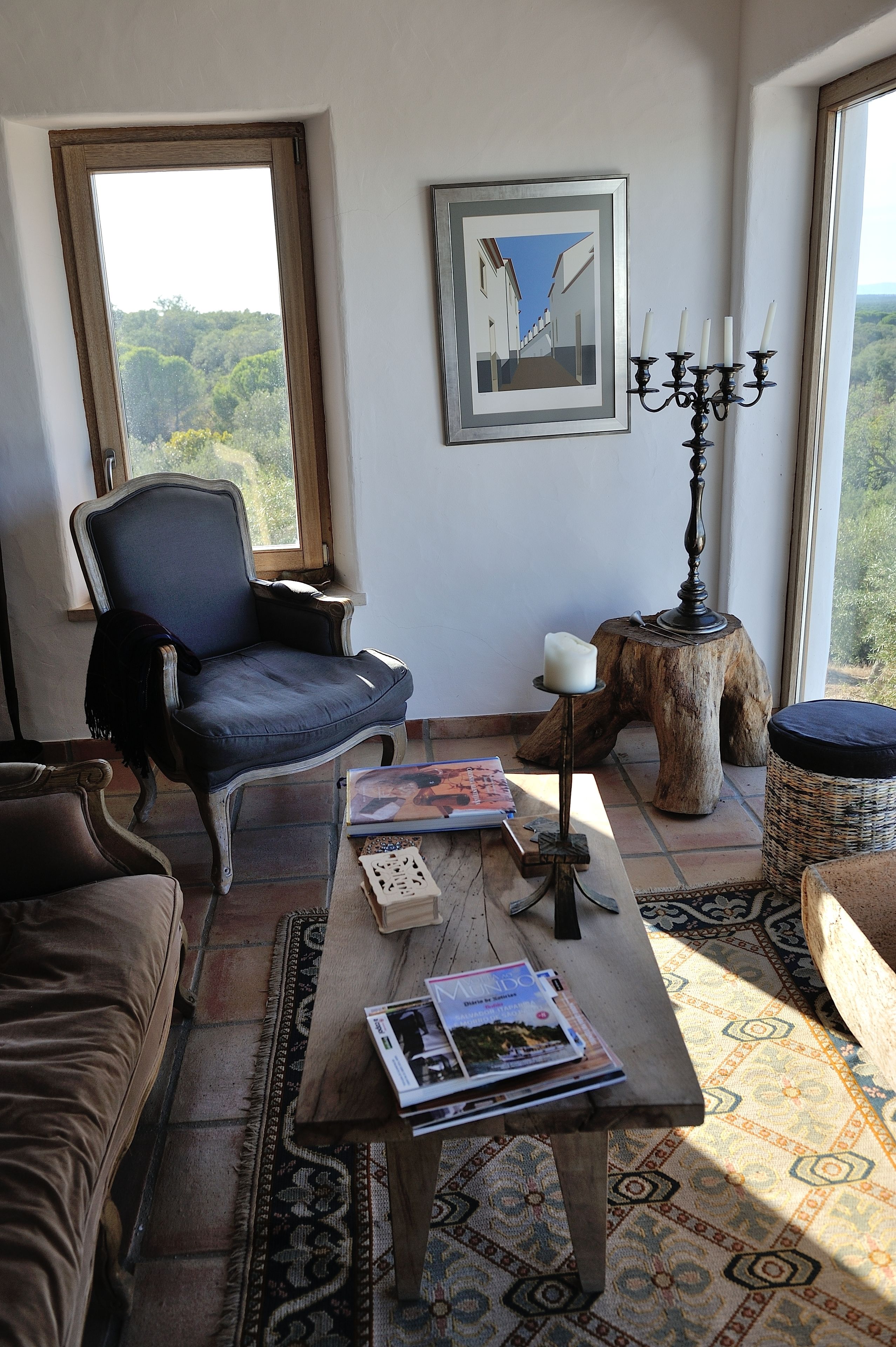 A cozy living room with a large window, a blue armchair, a wooden coffee table with magazines, and a candelabra.