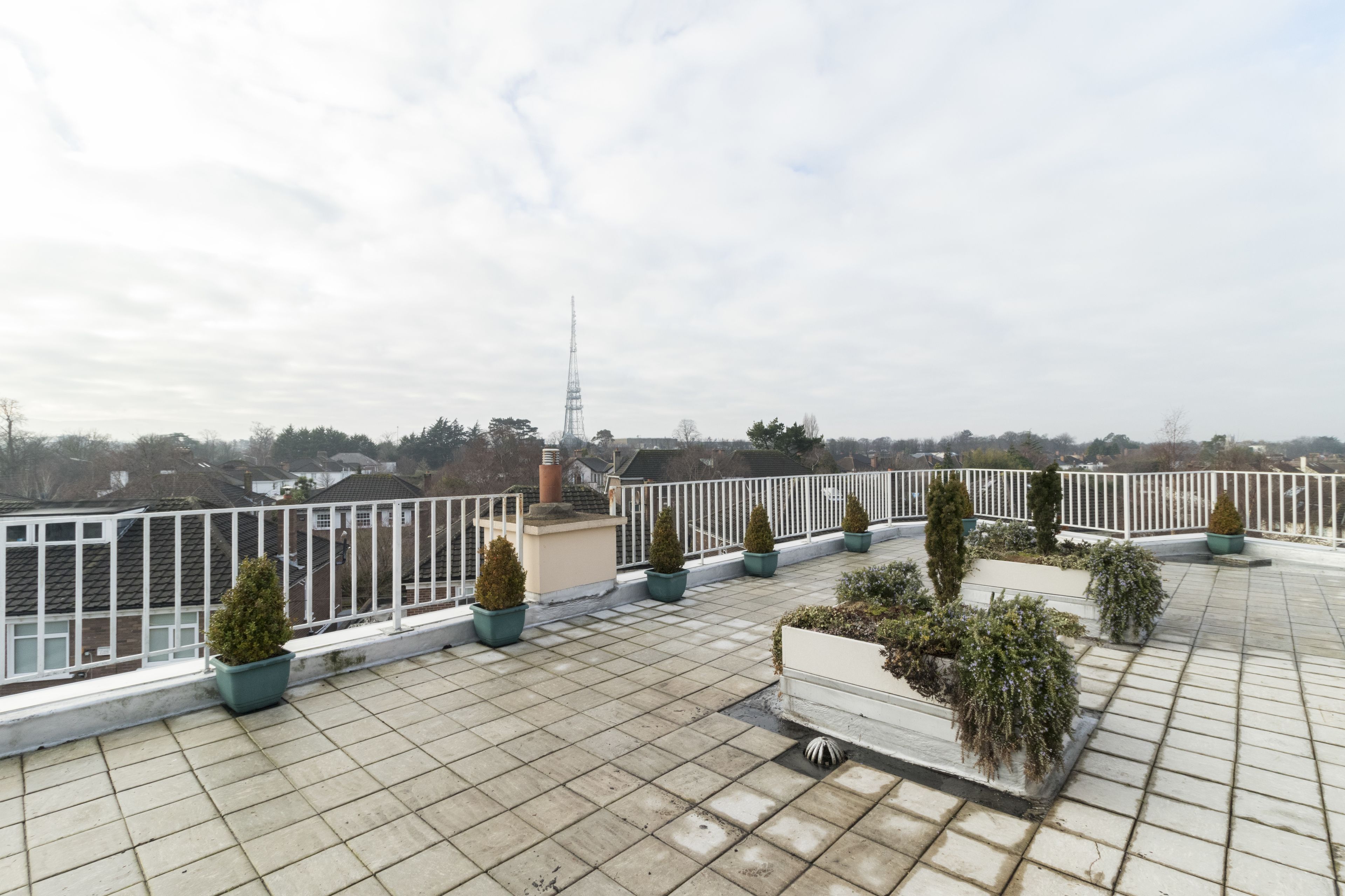 Een terras op het dak, met betegelde vloer, planten in potten en een witte balustrade.