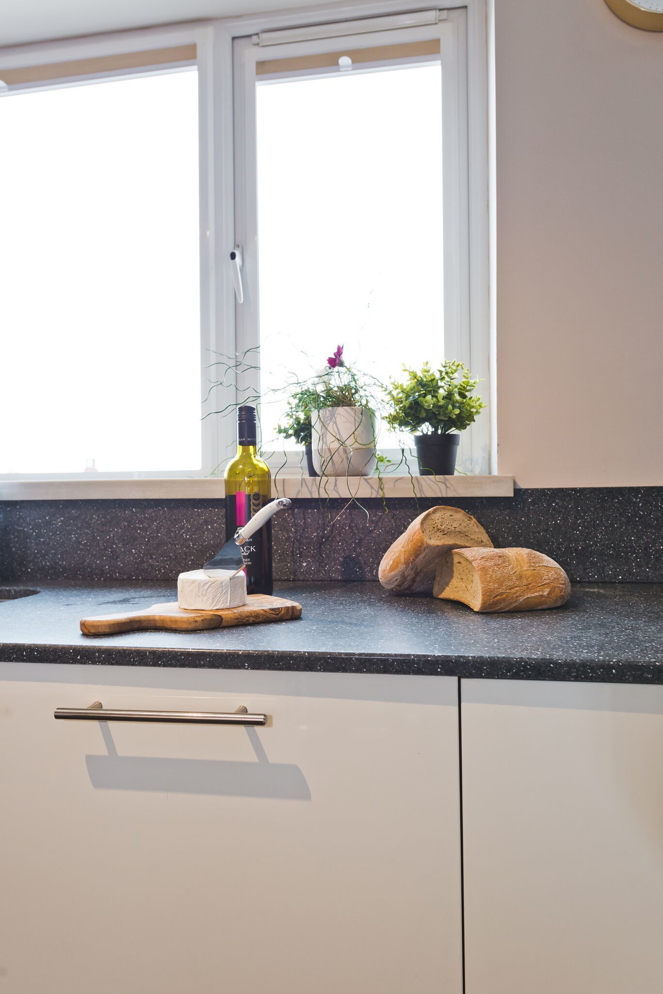 A kitchen counter with a bottle of wine, a cheese wheel, and two loaves of bread.
