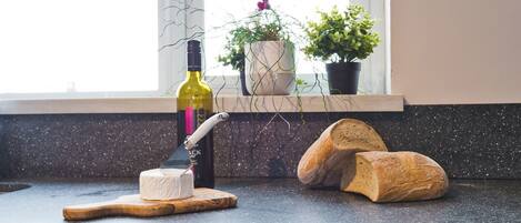 A kitchen counter with a bottle of wine, a cheese wheel, and two loaves of bread.