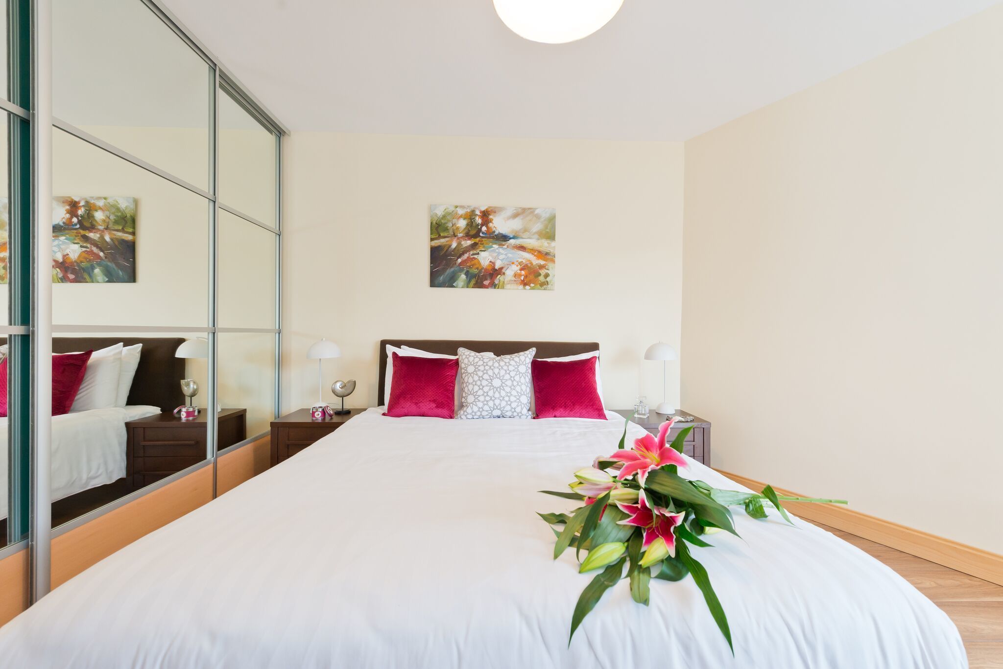 A hotel room with a large bed, a mirror, artwork on the wall, and a flower arrangement.
