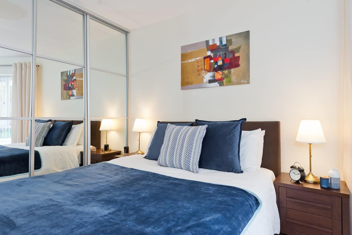 A modern hotel room with a large bed, a nightstand with a lamp, a clock, and a mirror.