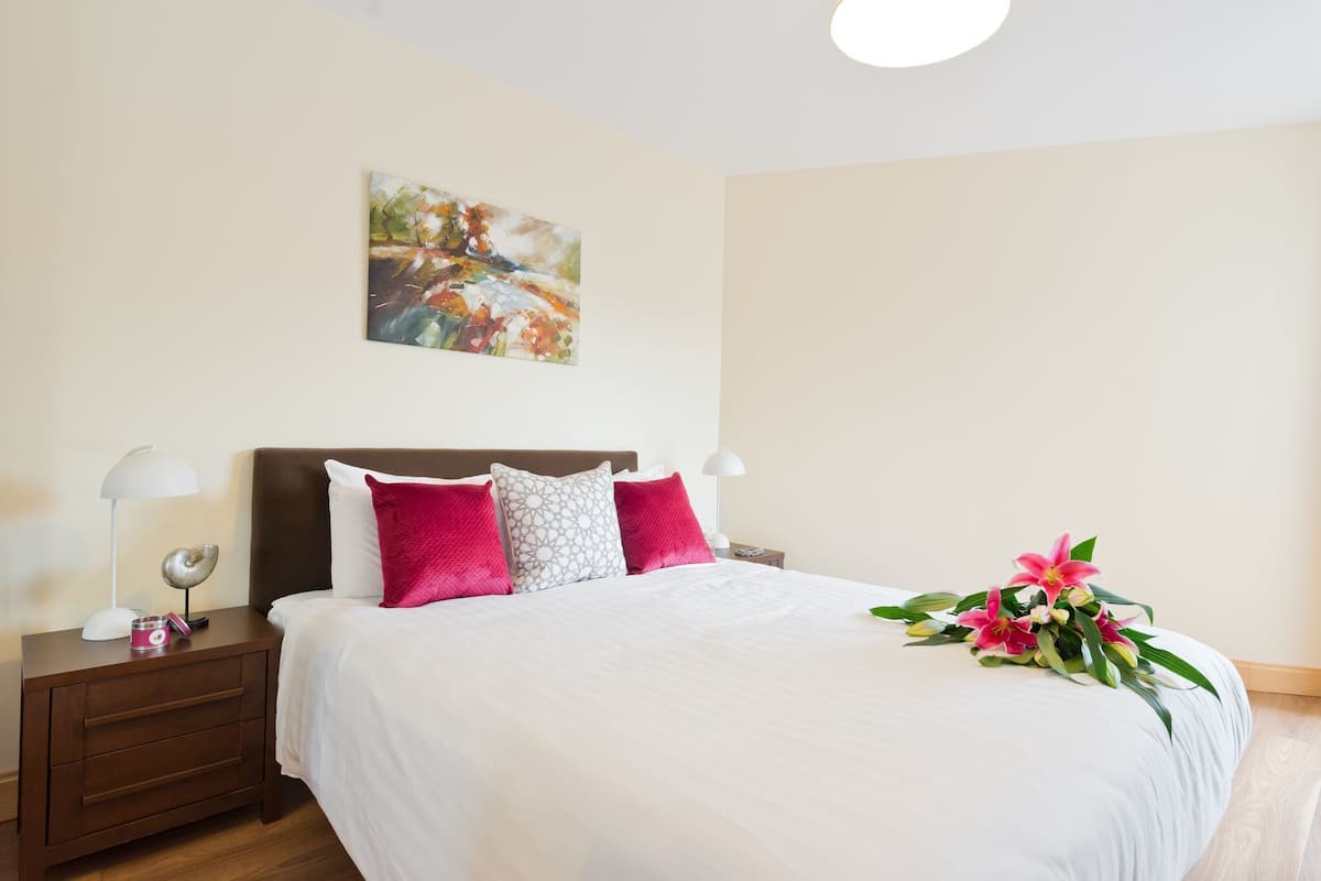 A neatly made bed with white and pink pillows, a bedside table with a lamp, and a painting on the wall.
