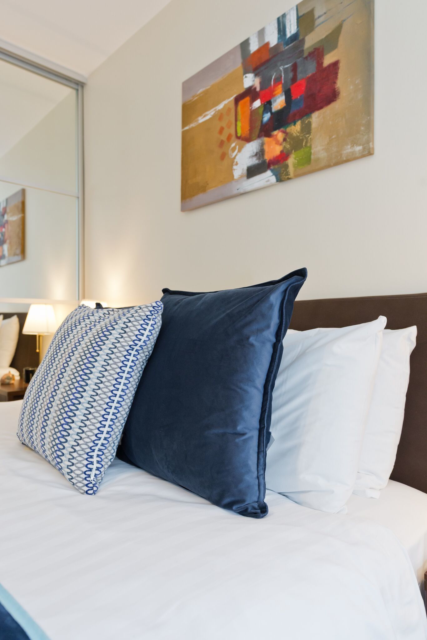 A neatly made bed with a patterned pillow, a solid blue pillow, and a white pillow. A colorful abstract painting hangs on the wall above the bed.