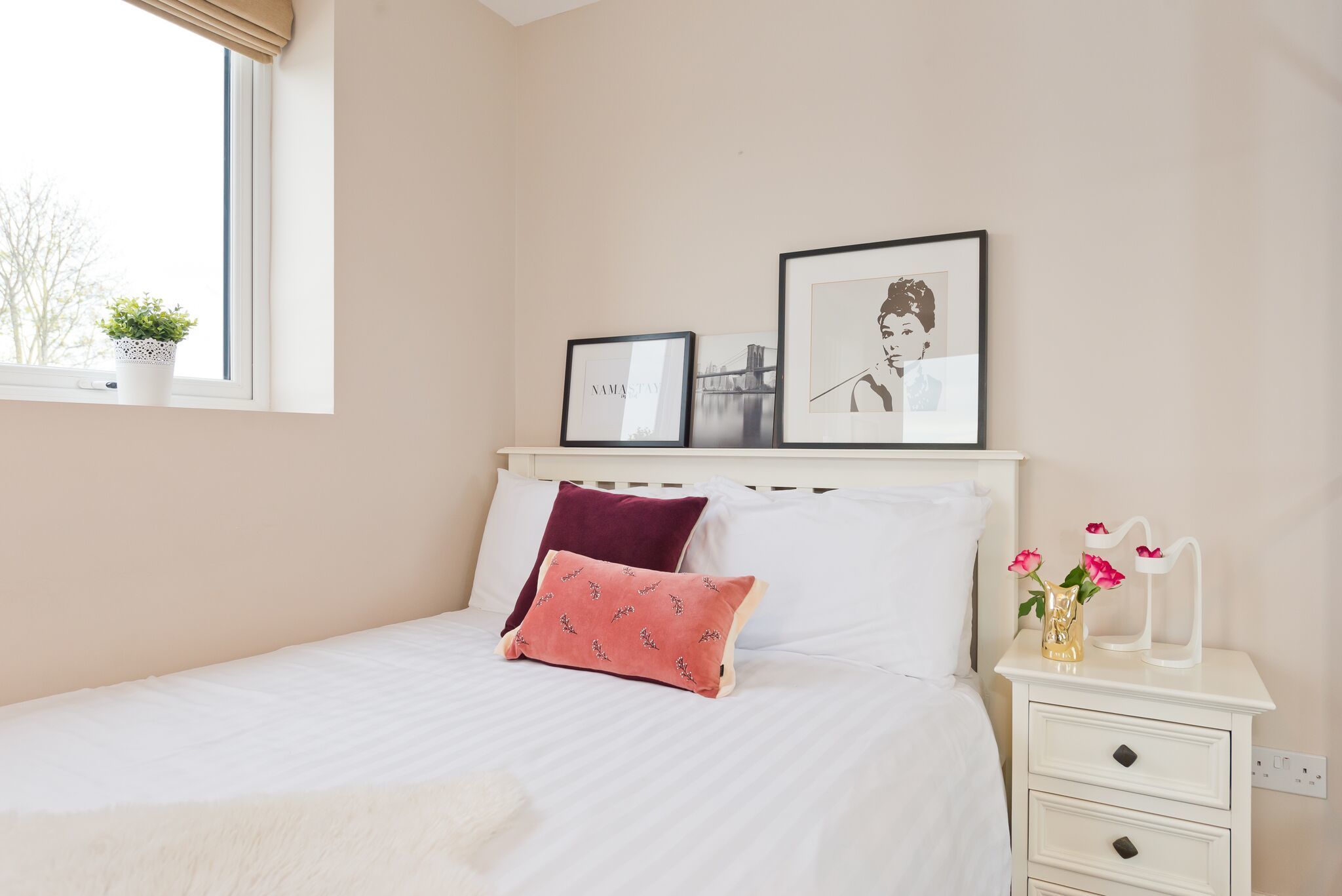 A bedroom with a bed, pillows, a nightstand with a vase of flowers, and framed pictures on the wall.