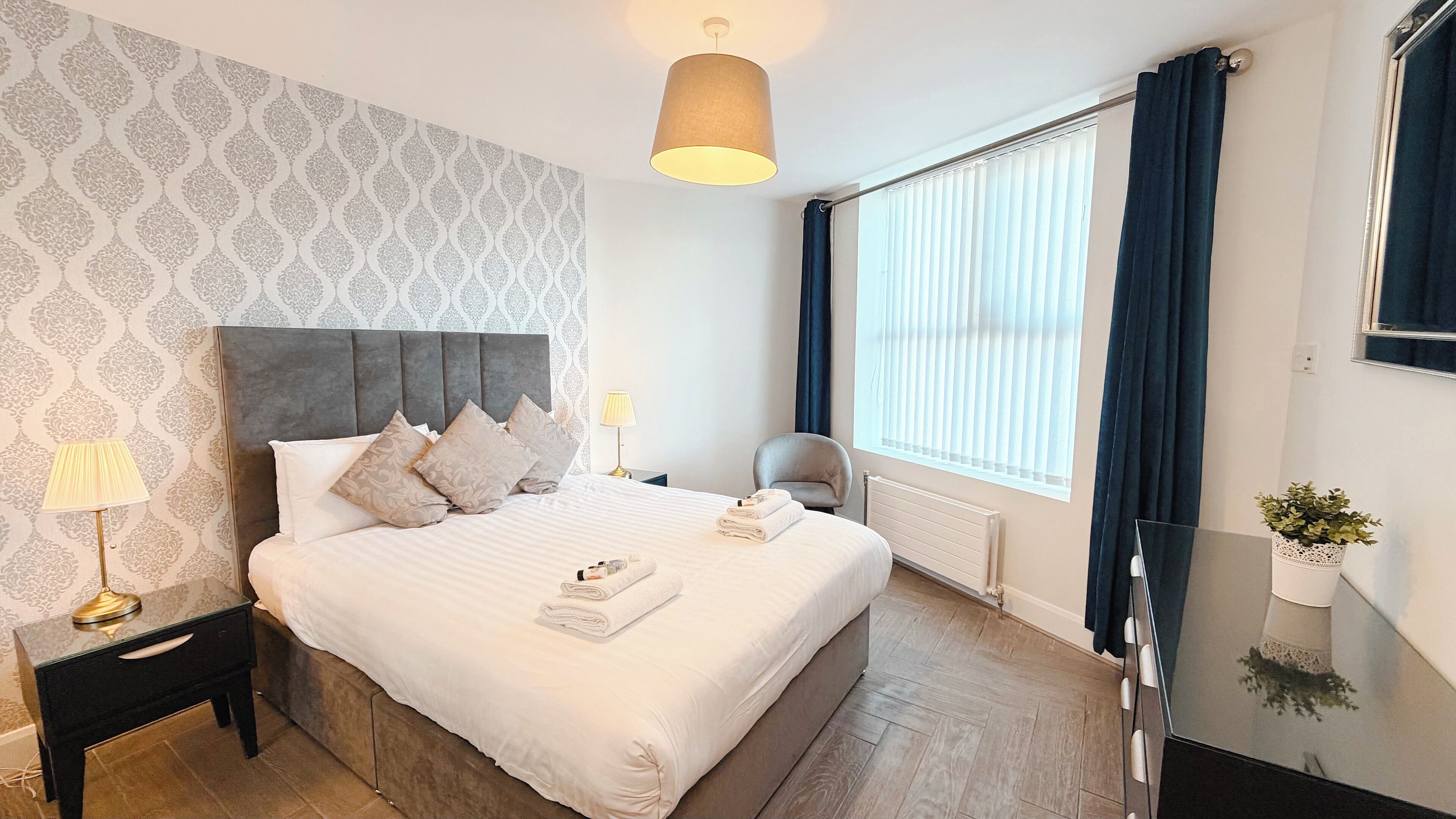 A neatly arranged hotel room with a large bed, a bedside table with a lamp, a radiator, a window with curtains, and a small potted plant.