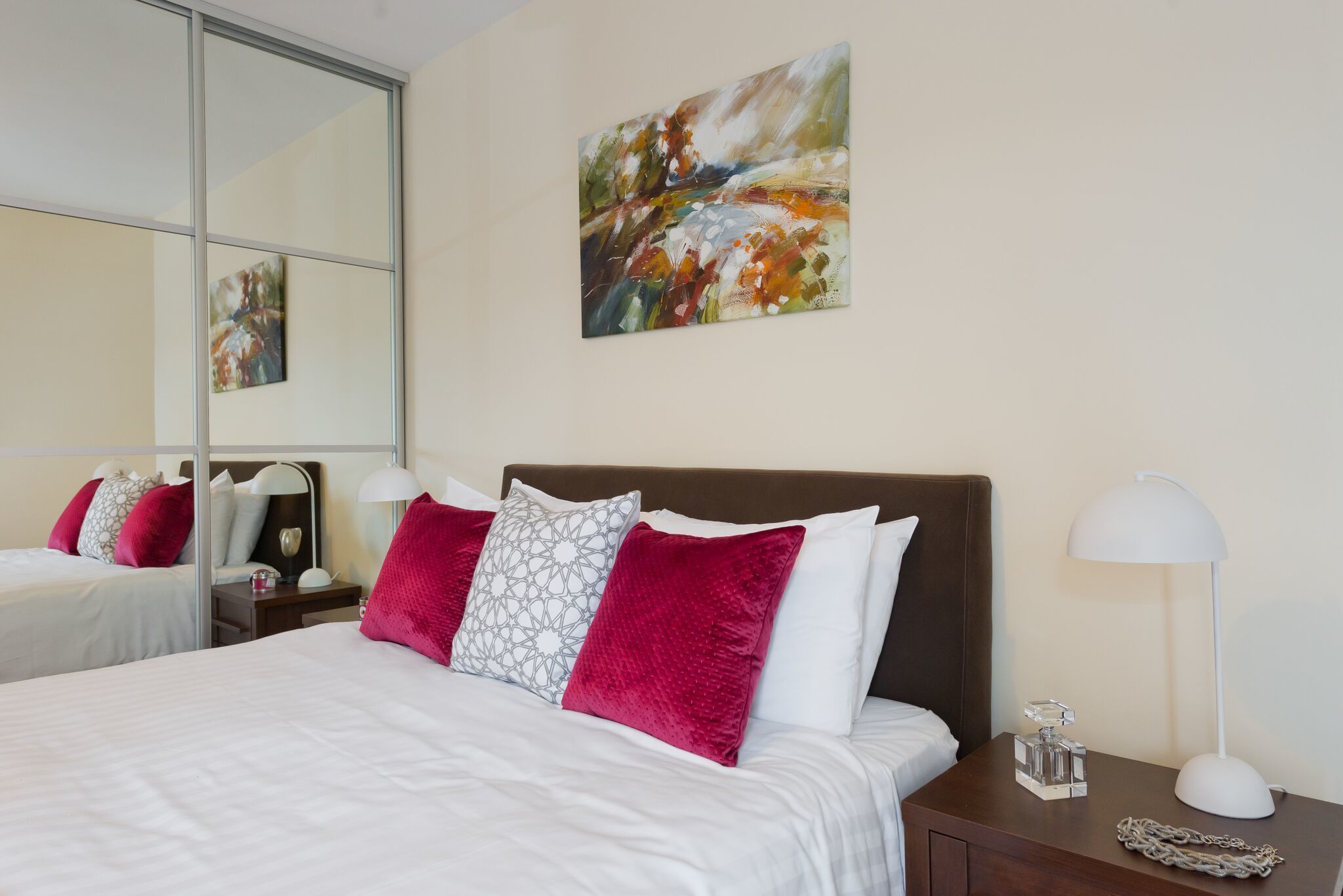 Habitación de hotel con una cama grande, ropa de cama blanca, almohadas rojas y estampadas, una mesita de noche con una lámpara y un cuadro en la pared.