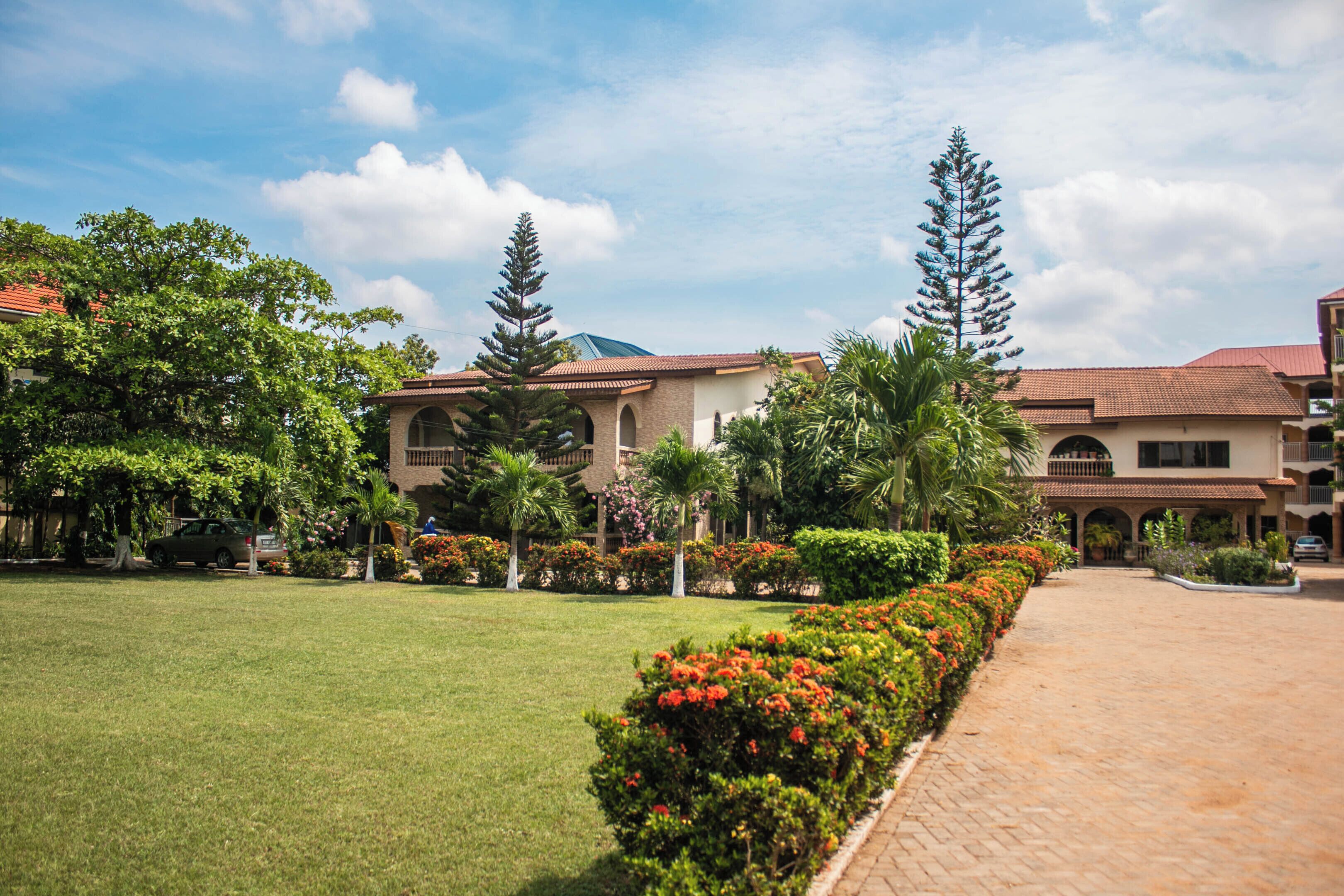 A courtyard with a paved walkway, surrounded by lush greenery and a building with a tiled roof.