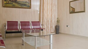 A waiting area with a glass table, red chairs, a painting, a floral curtain, and a mirror on the wall. - Sheridan Hotel (Accra)