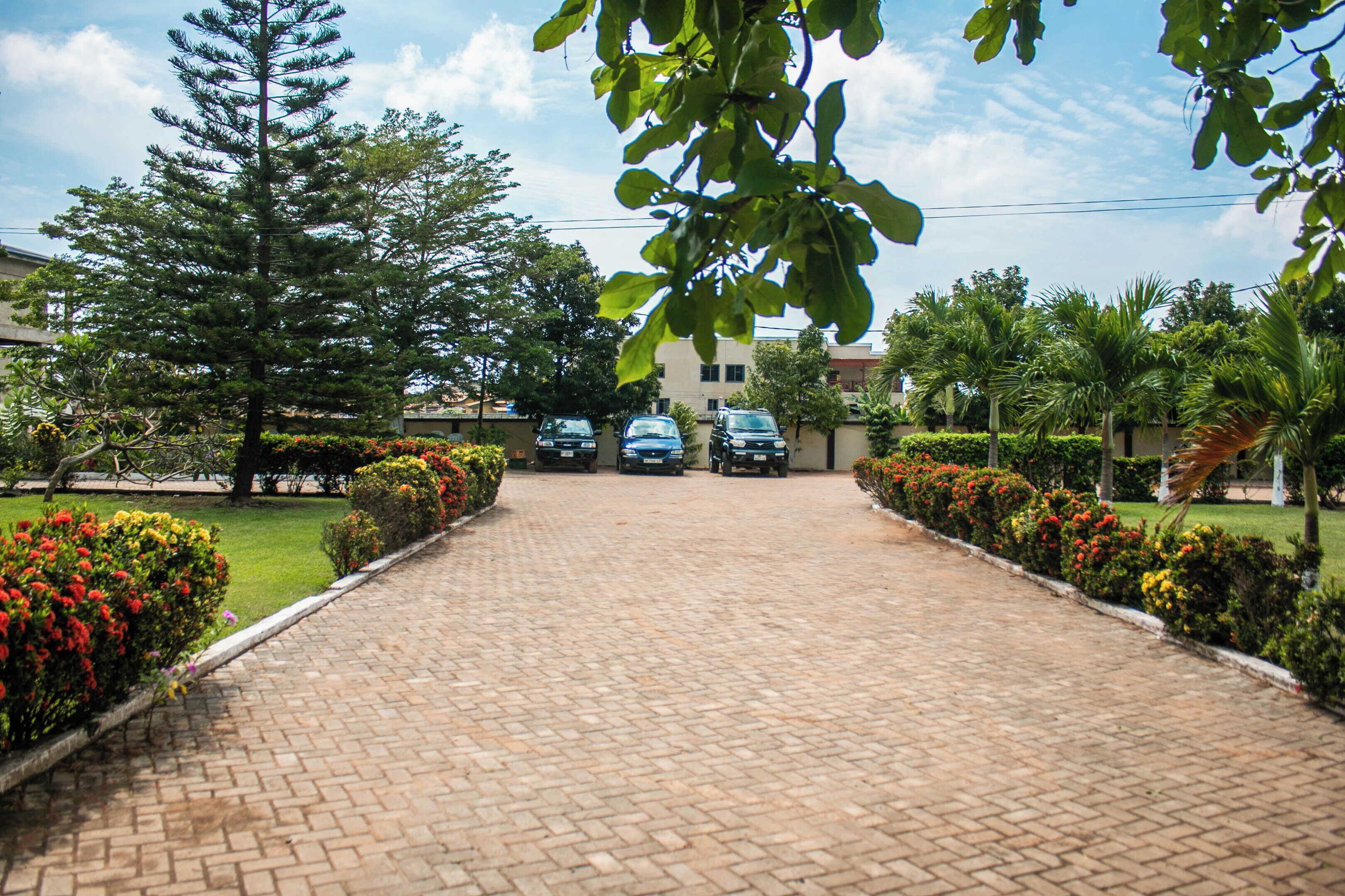 A paved driveway leading to a building with parked cars, surrounded by well-maintained gardens and trees.