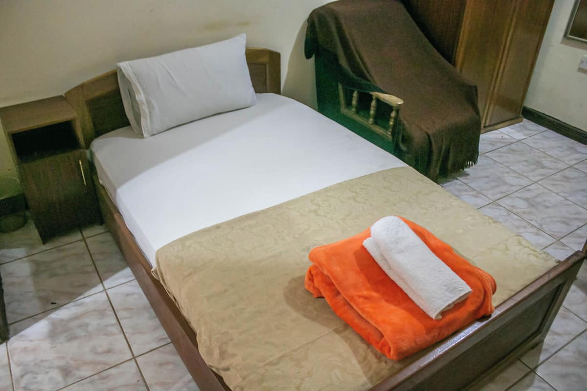A single bed with a white pillow, an orange blanket, and a folded white towel on top.