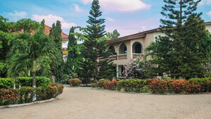 A paved courtyard with a building featuring arched windows and a balcony, surrounded by lush greenery and flowering plants. - Sheridan Hotel (Accra)