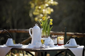 Une table dressée avec une théière blanche, des tasses, un bol et des pots de confiture, avec des jonquilles jaunes en arrière-plan.