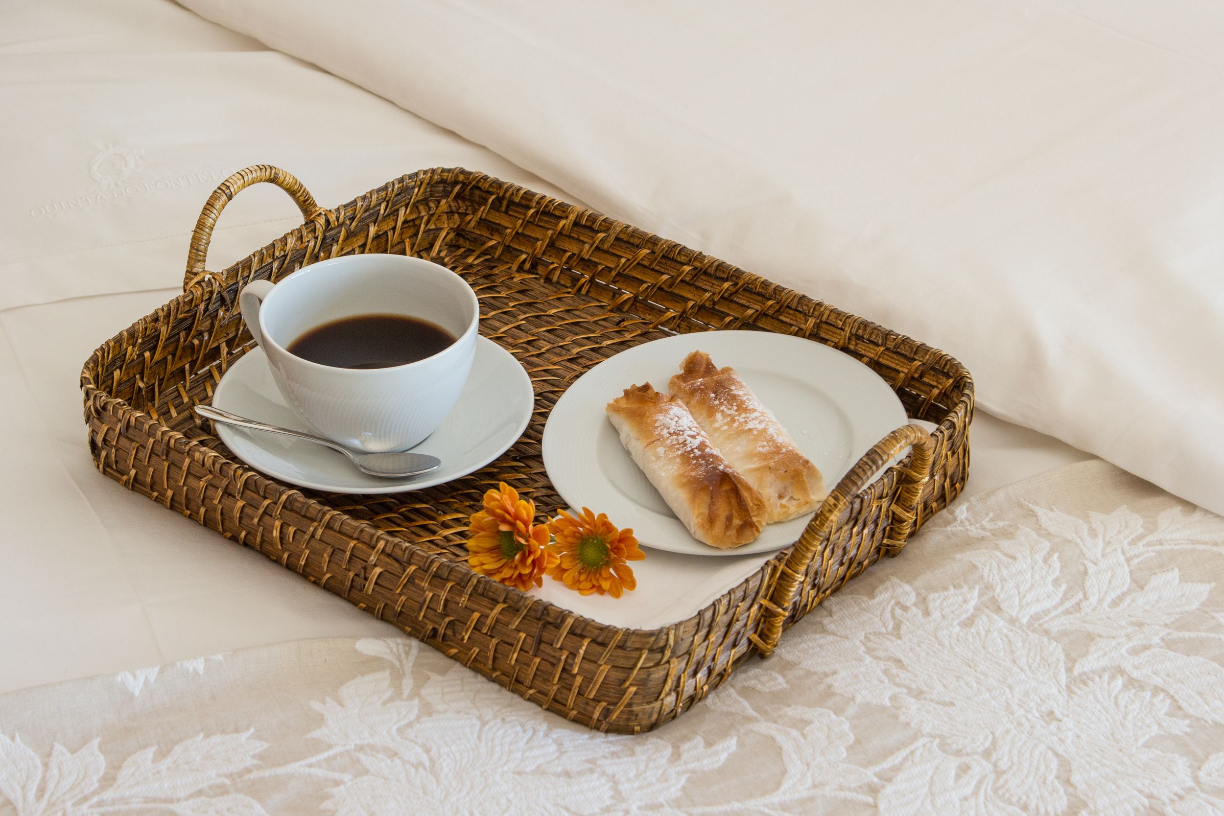 Una bandeja de mimbre con una taza de café, un plato con un pastel y una flor sobre una cama con una colcha blanca.