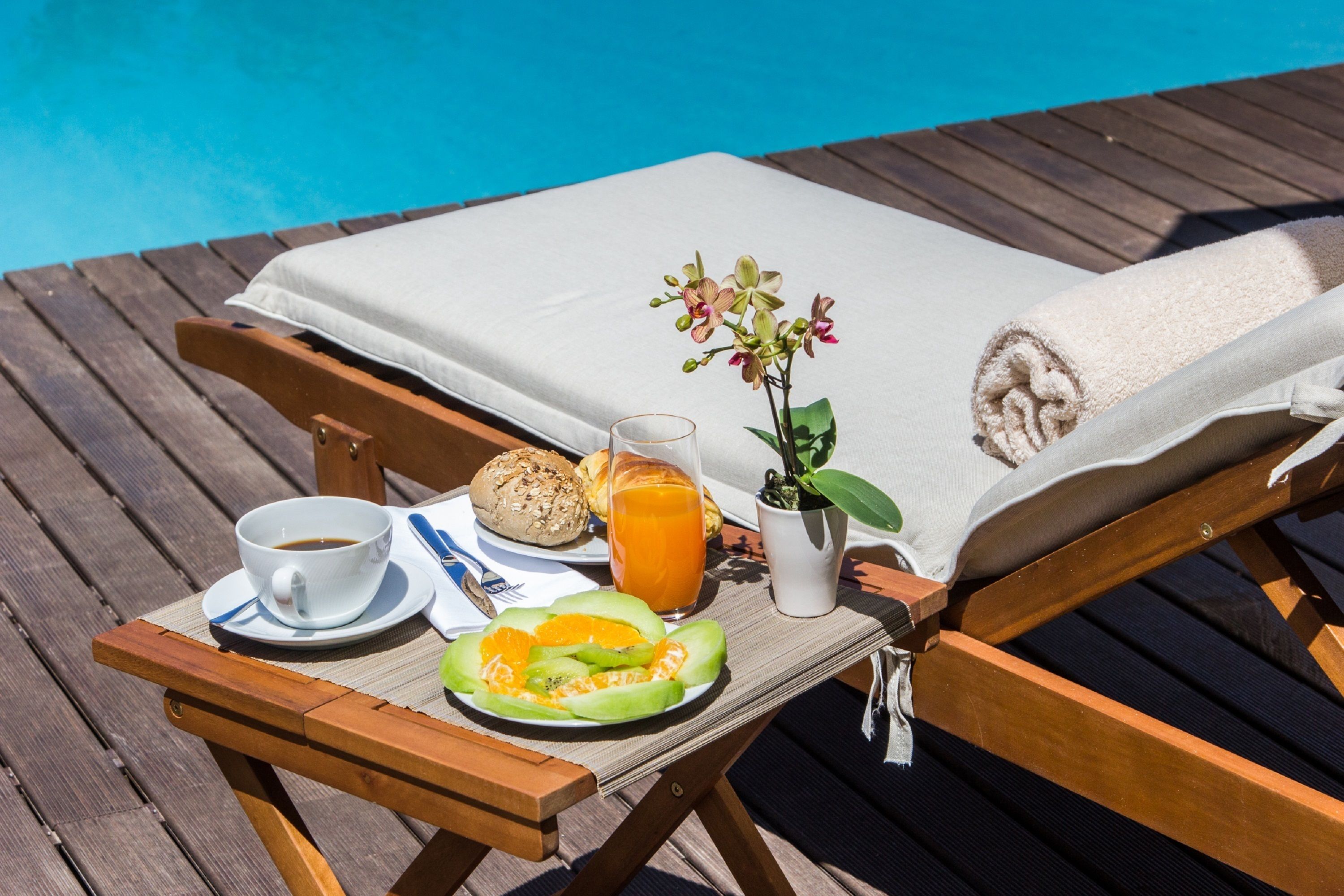 A wooden deck with a lounge chair, a table with breakfast items including a cup of coffee, a glass of orange juice, a plate with melon slices, and a small potted plant.