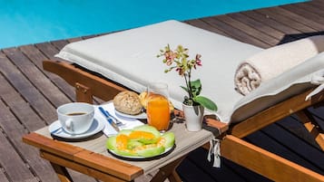 A wooden deck with a lounge chair, a table with breakfast items including a cup of coffee, a glass of orange juice, a plate with melon slices, and a small potted plant.