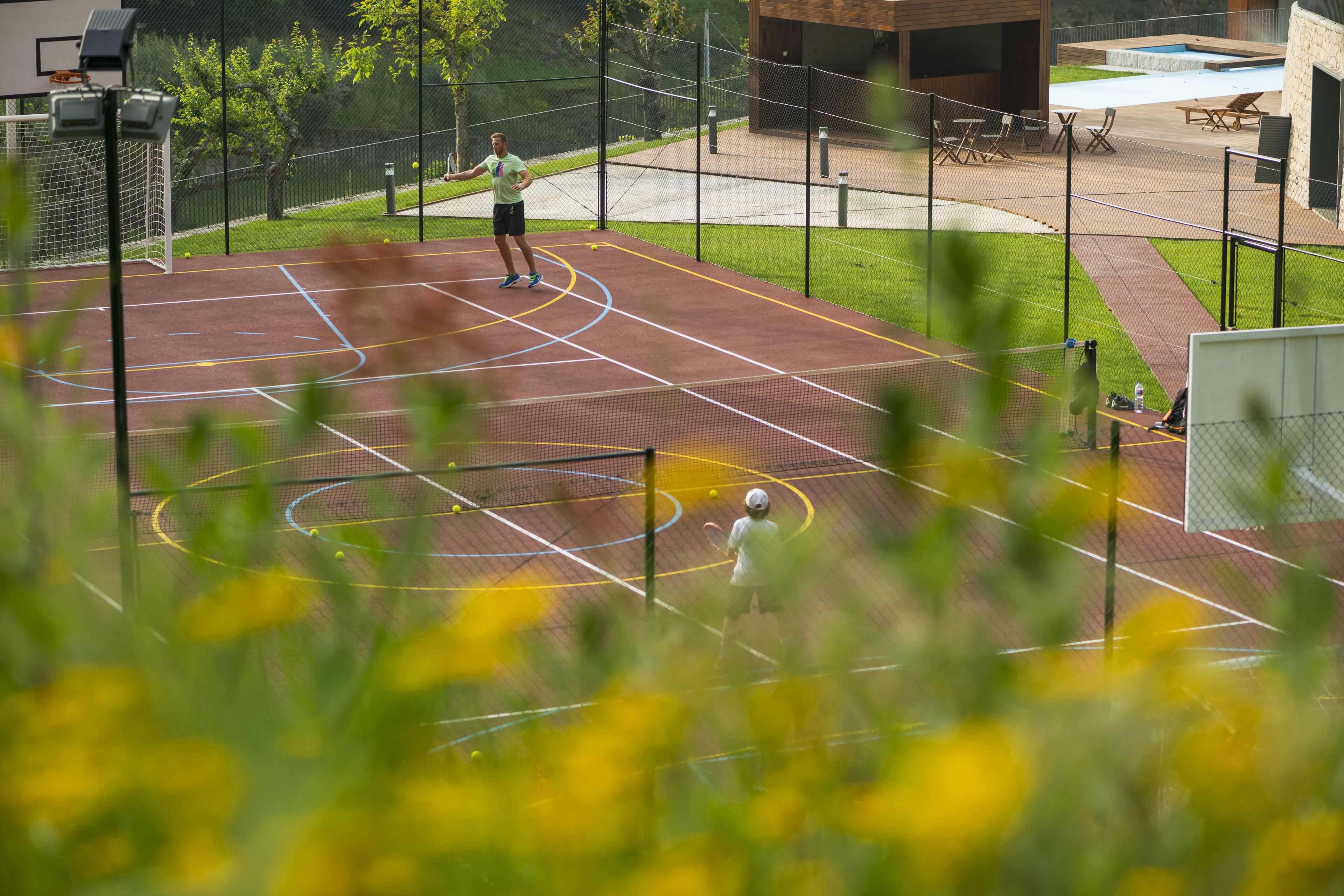 Una cancha de tenis al aire libre con una red, un aro de baloncesto y un pequeño espacio de descanso con sillas.
