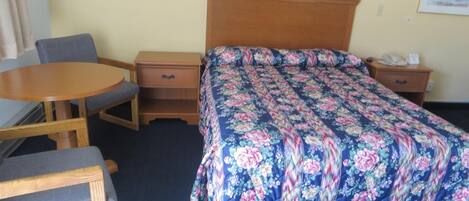 A hotel room with a bed, a wooden nightstand, a desk with a chair, and a small round table.
