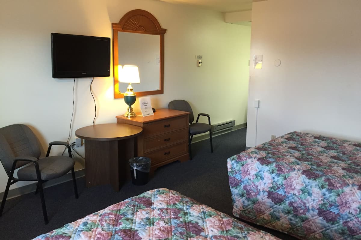 A hotel room with two single beds, a desk with a lamp, a chair, a television mounted on the wall, and a mirror above the desk.