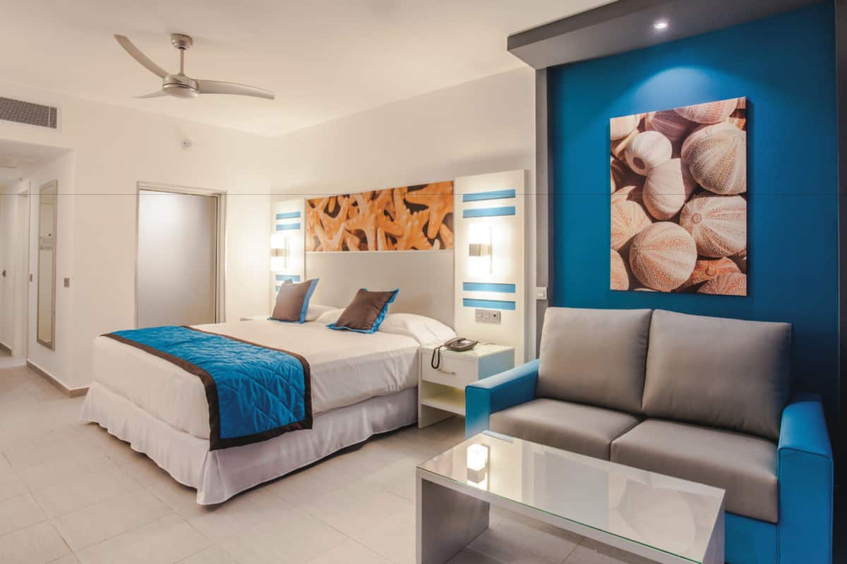 A modern hotel room with a bed, a sofa, a glass coffee table, a ceiling fan, and a wall with a decorative painting.