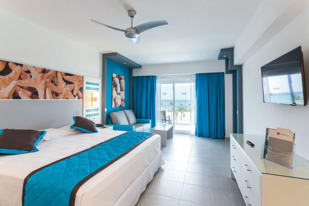 A modern hotel room with a bed, a desk, a chair, a ceiling fan, and a TV.