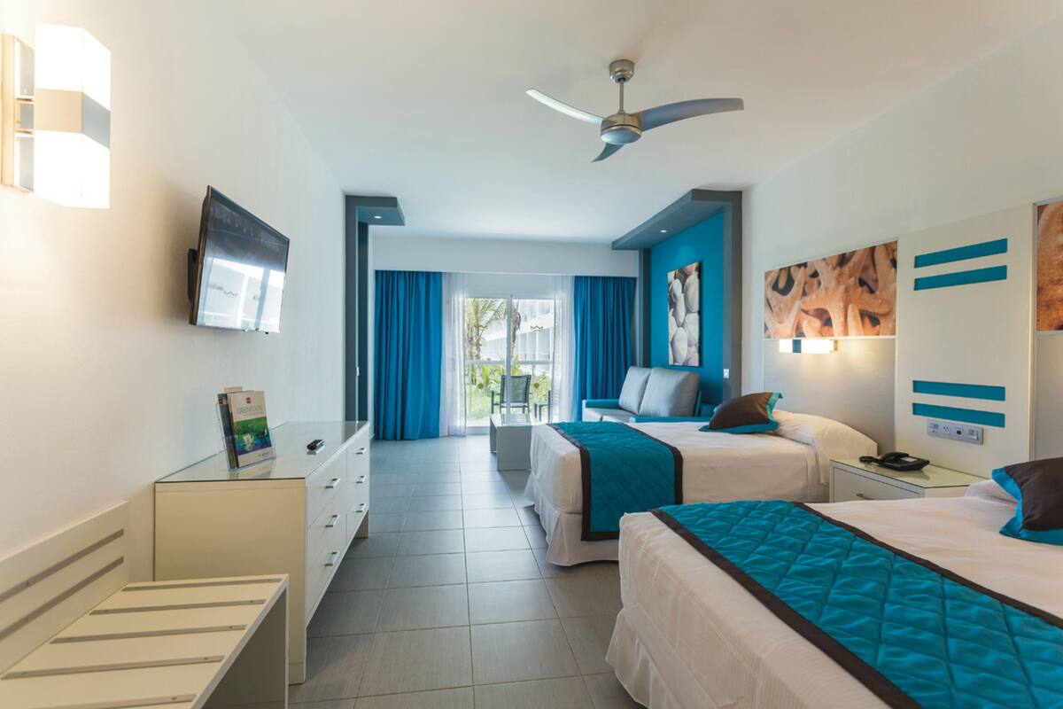 A hotel room with two beds, a ceiling fan, a TV, and a view of the outdoors.