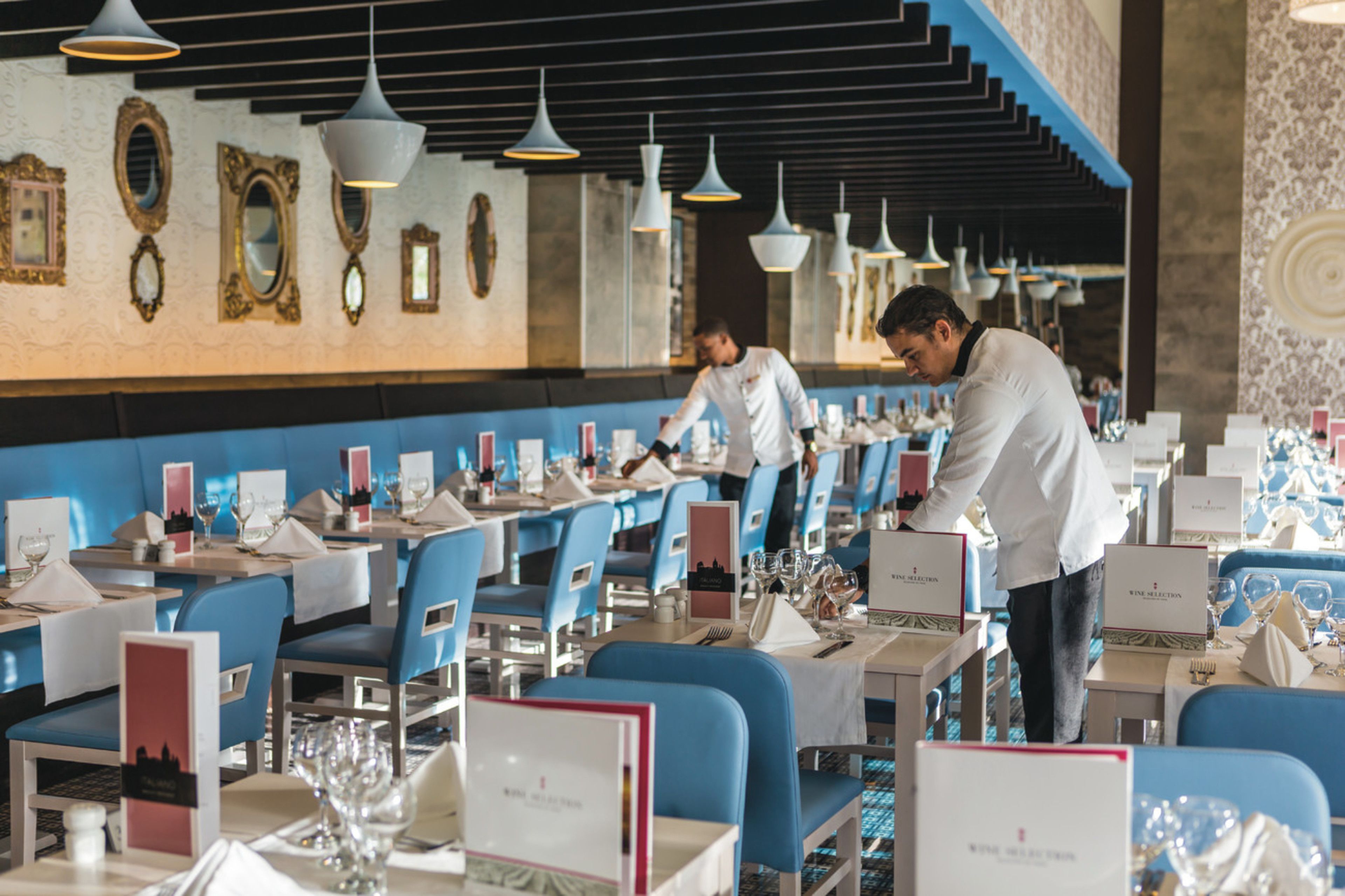 Un restaurant avec des chaises et des tables bleues, un chef qui prépare les plats et un serveur qui apporte les commandes.