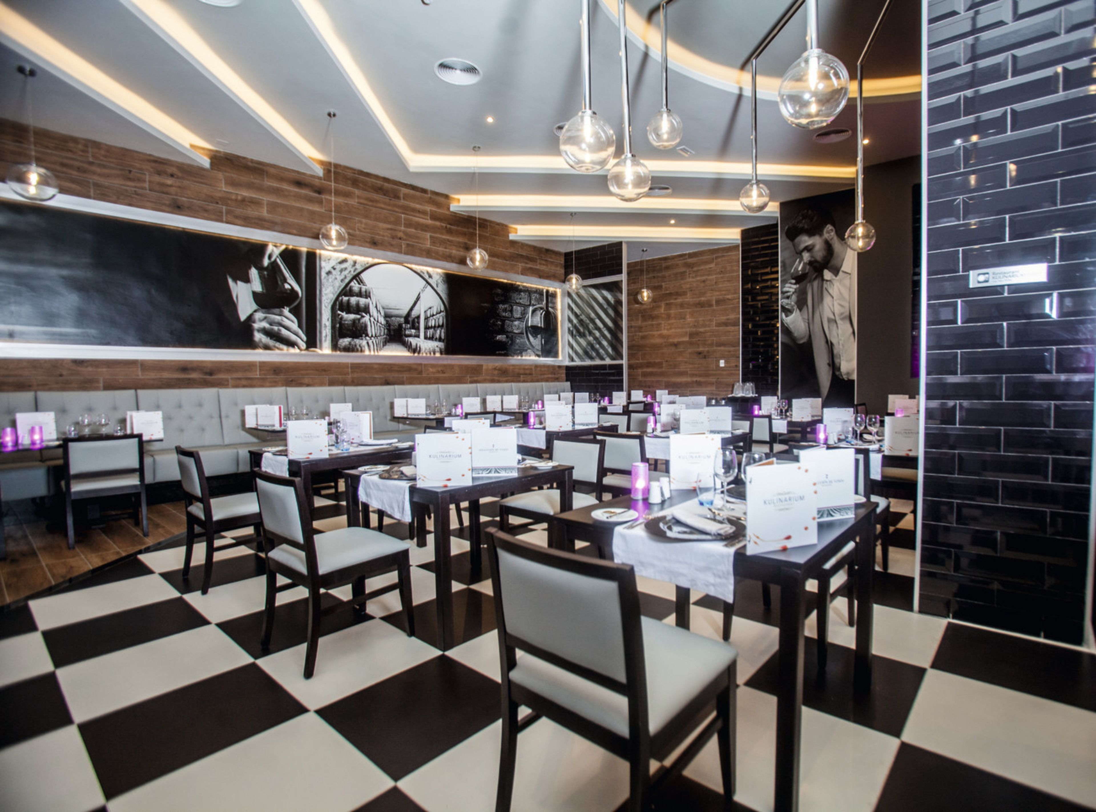 A restaurant with a checkered floor, wooden walls, and hanging spherical light fixtures. Tables are set with white tablecloths and menus.