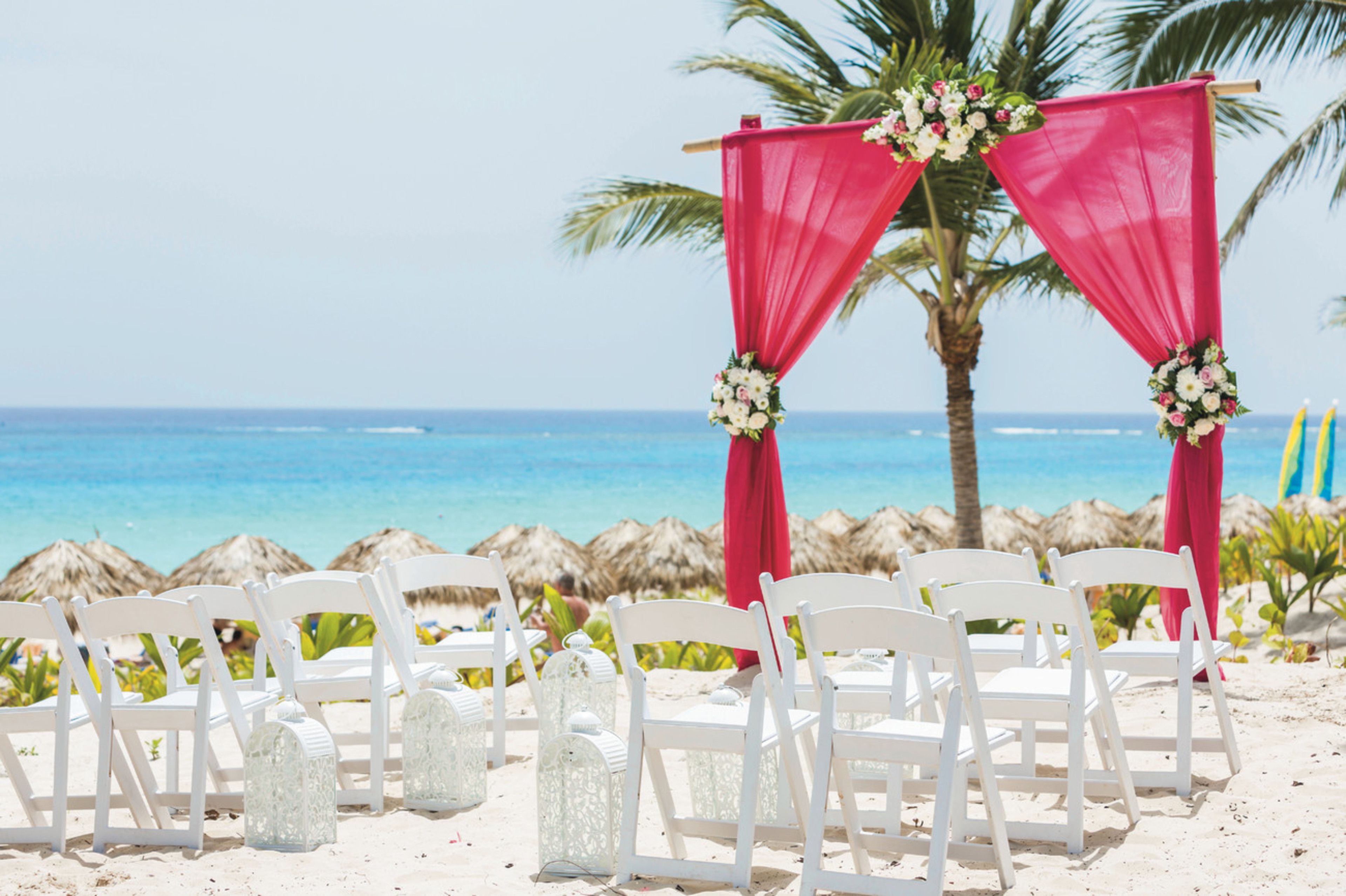 Une cérémonie de mariage organisée sur la plage, avec des chaises blanches et des draperies rouges, offrant une vue sur l’océan.
