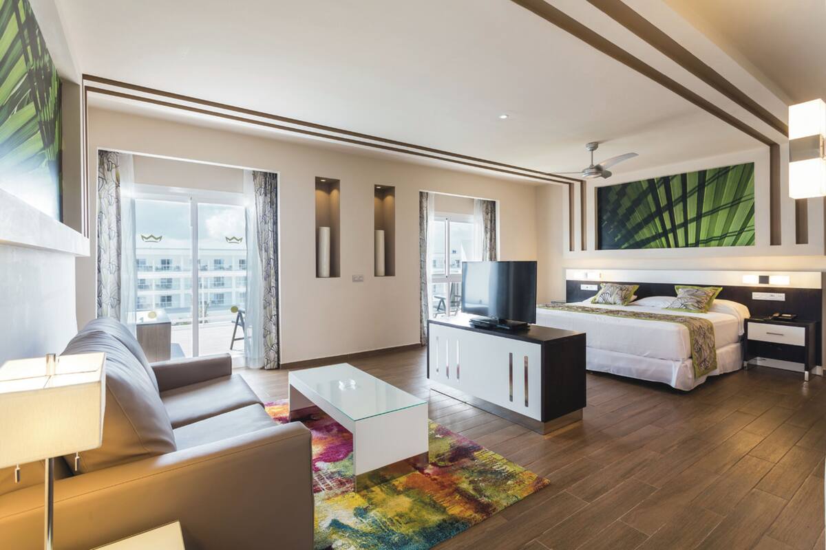 A modern hotel room with a sofa, a glass coffee table, a bed with a TV, and a large window with curtains.