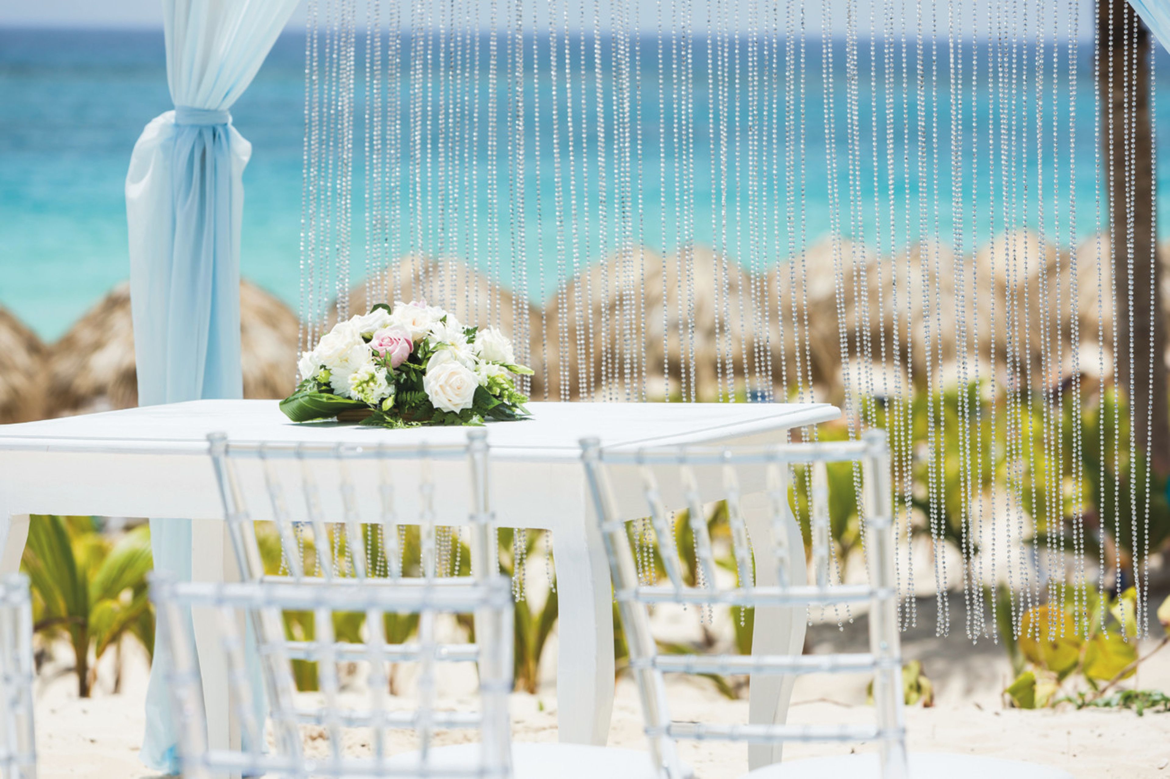 A white table with chairs set for two, positioned on a sandy beach with a view of the ocean and rocks in the background.