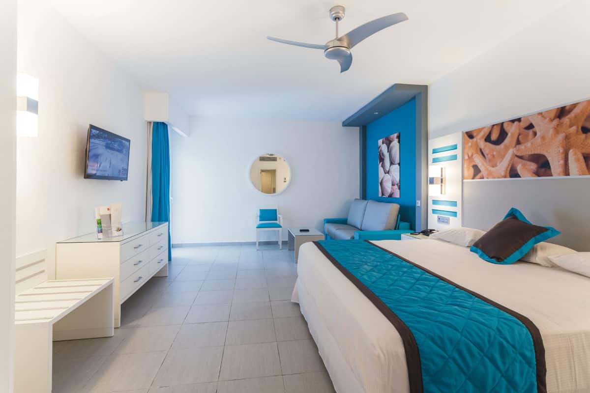 A modern hotel room with a bed, a desk, a chair, a TV, and a ceiling fan.