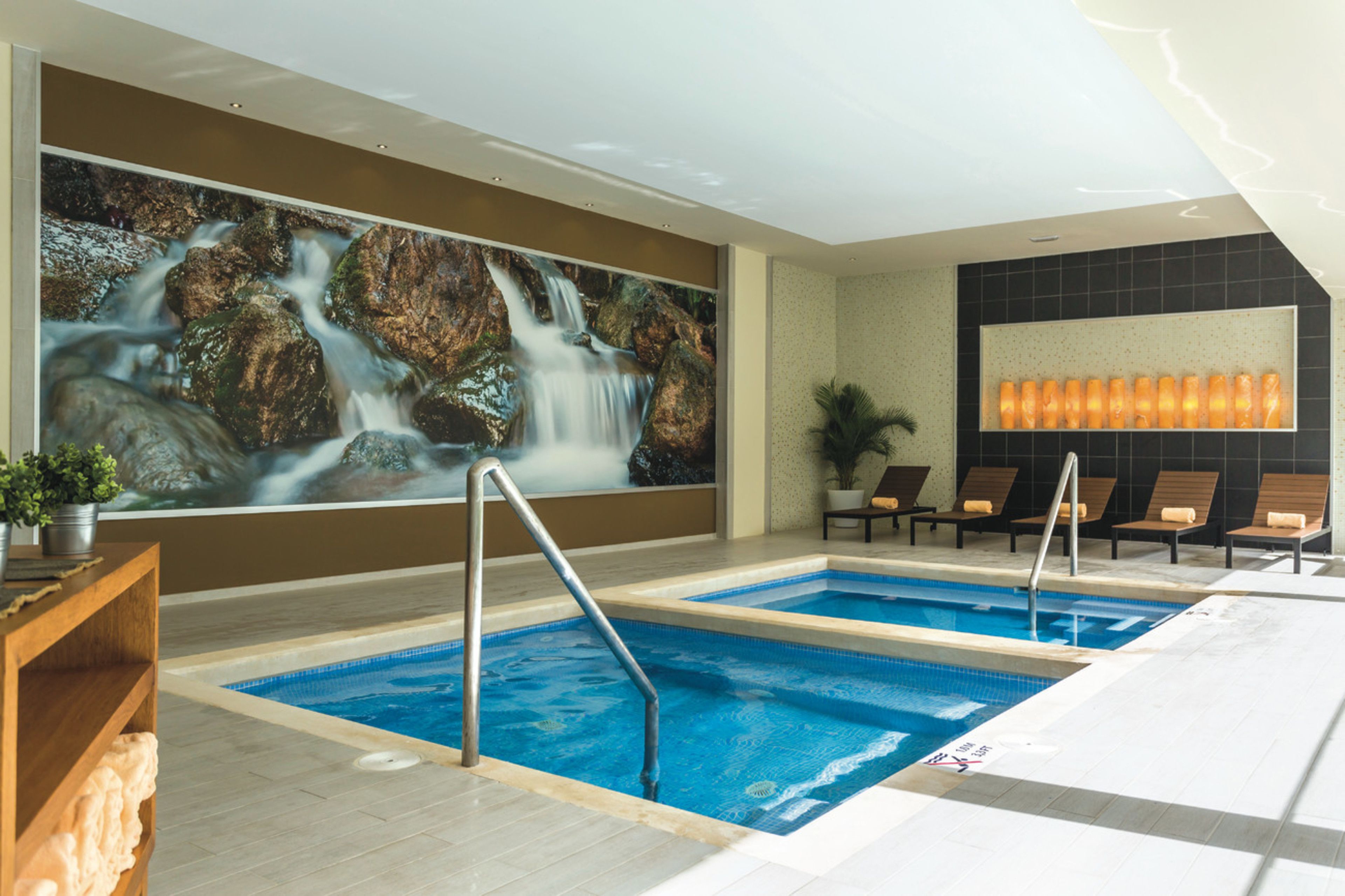 Ein Hotelpoolbereich mit zwei Pools, einer Wandmalerei mit einem Wasserfall, Liegestühlen und einer Wand mit einer Leuchte.