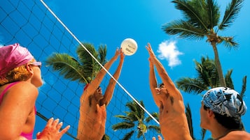 Un match de volleyball de plage, avec des joueurs qui sautent pour frapper la balle, des palmiers et un ciel bleu dégagé.