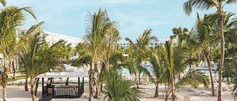 A beach area with palm trees, a covered seating area, and a pool in the background.