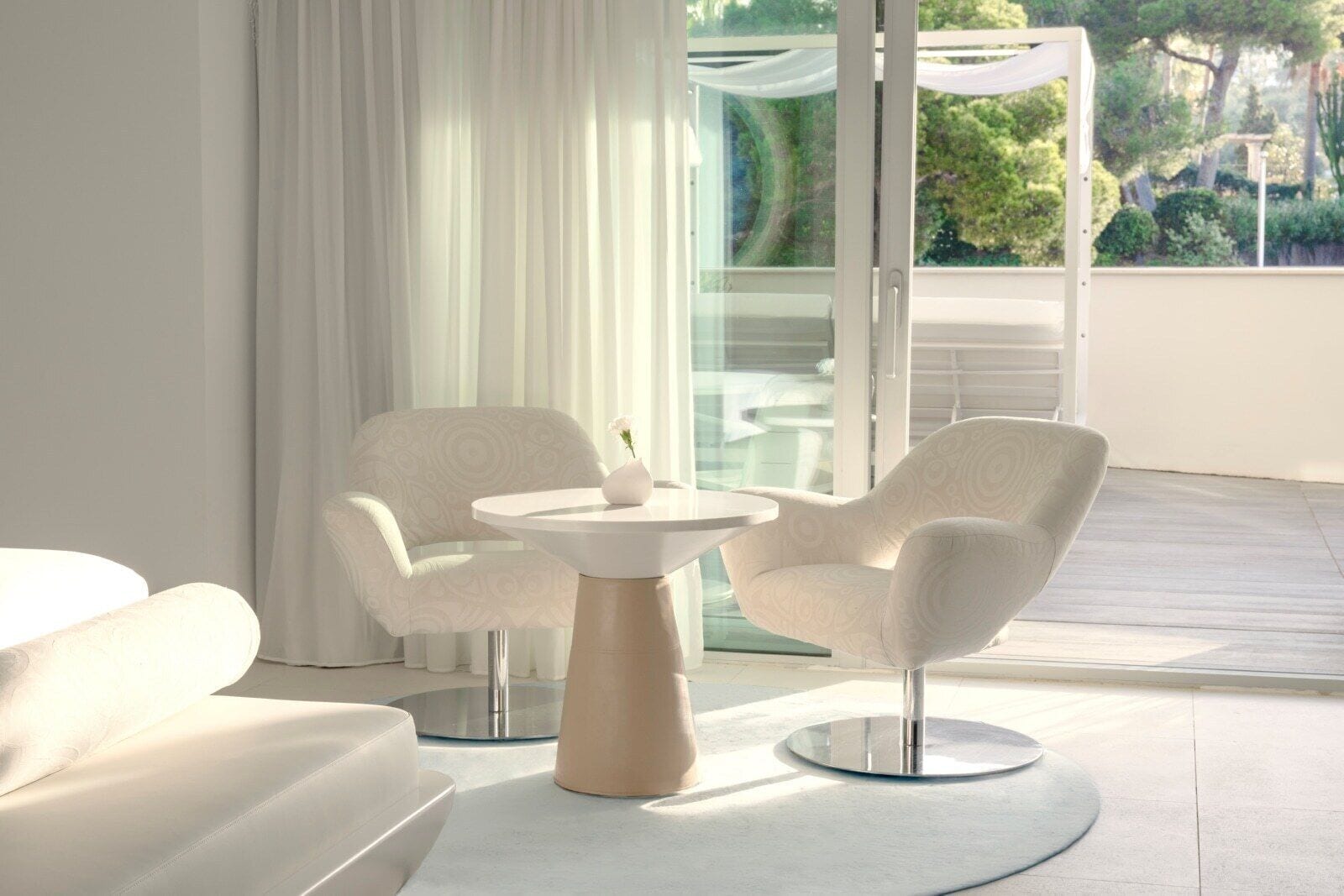 Habitación moderna y minimalista con dos sillones blancos y una mesita, una ventana grande con cortinas translúcidas y vistas a espacios verdes.