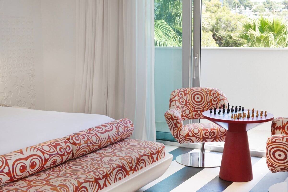 A hotel room with a bed, a patterned sofa, a red table with chess pieces, and a view of greenery outside.