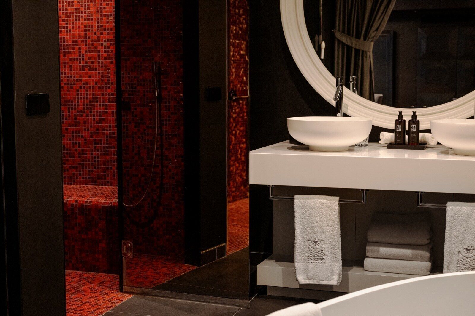 A modern bathroom with a large mirror, two sinks, and a walk-in shower with red mosaic tiles.