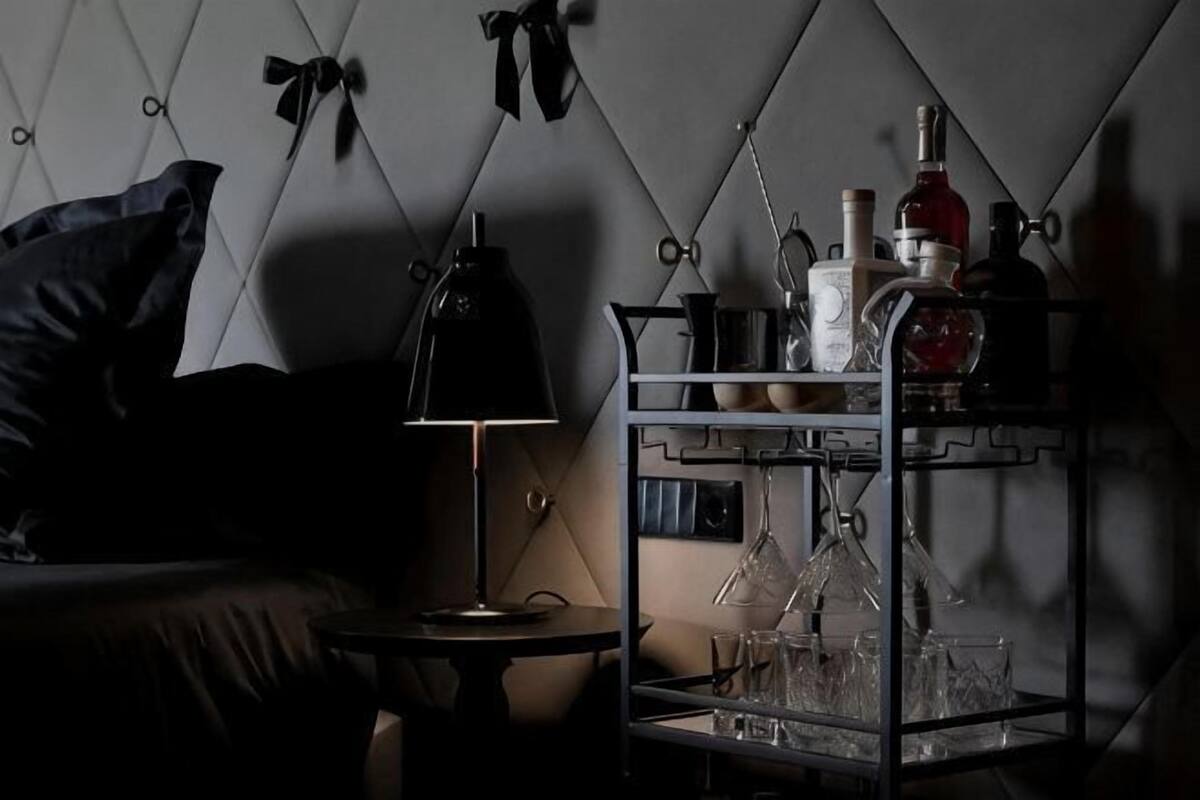 A dimly lit room with a bed, a small table with a lamp, and a metal shelf with bottles and glasses.
