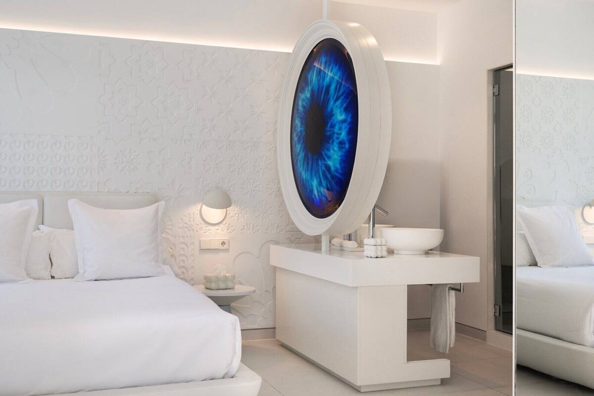 A modern hotel room with a large bed, a round mirror, a white vanity with a sink, and a decorative wall panel.