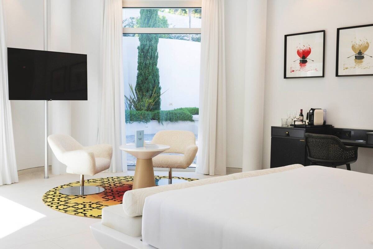 A hotel room with a bed, two chairs, a desk, a TV, and artwork on the wall.